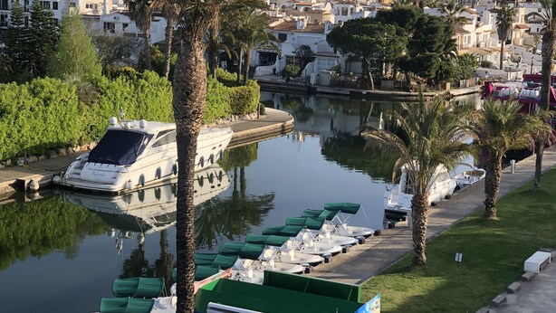 A marina with boats docked, a large yacht, and a backdrop of residential buildings and mountains.