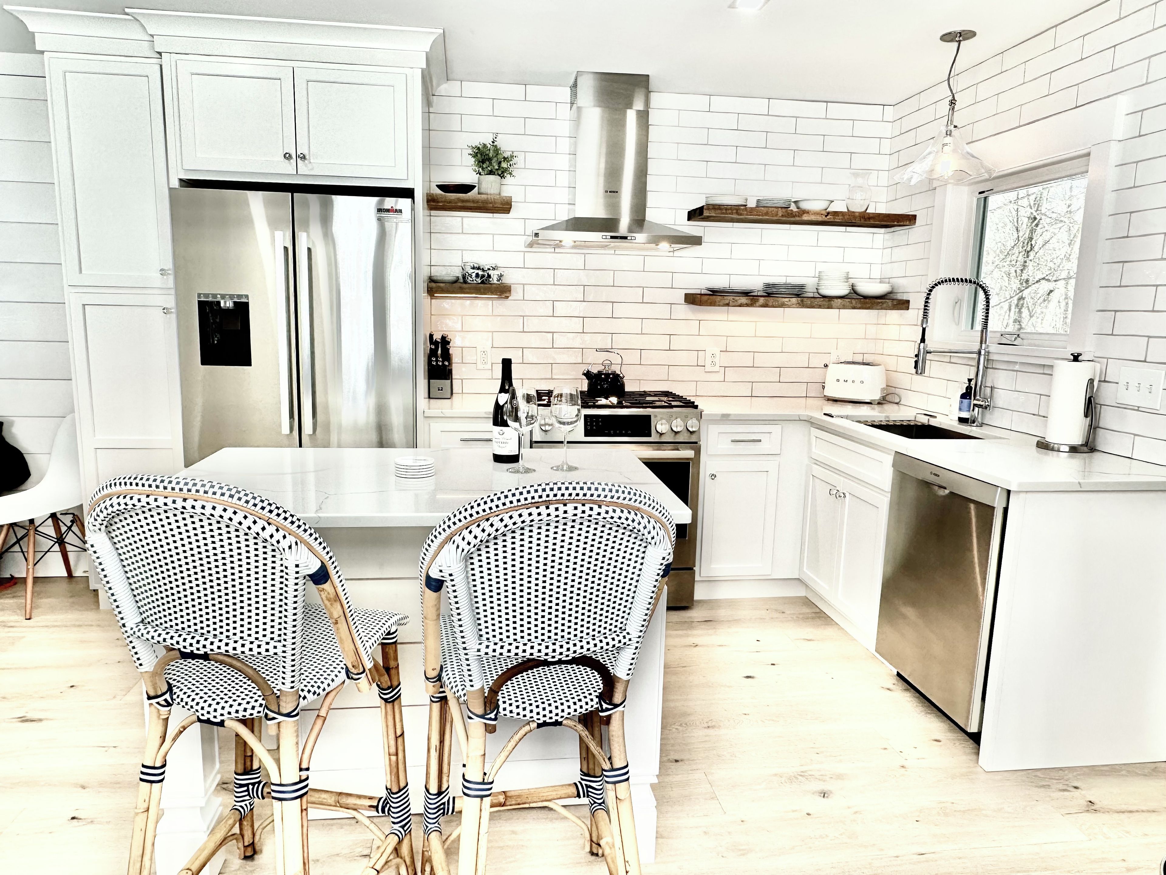 Una cocina moderna con armarios blancos, un refrigerador de acero inoxidable, una estufa a gas y un lavavajillas. Hay dos sillas de mimbre junto a una mesa blanca.