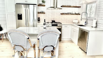 Una cocina moderna con armarios blancos, un refrigerador de acero inoxidable, una estufa a gas y un lavavajillas. Hay dos sillas de mimbre junto a una mesa blanca.
