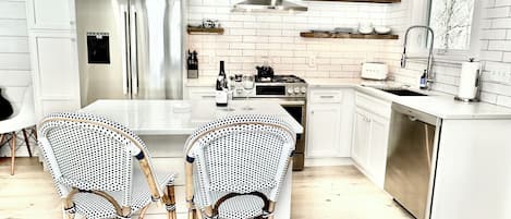Una cocina moderna con armarios blancos, un refrigerador de acero inoxidable, una estufa a gas y un lavavajillas. Hay dos sillas de mimbre junto a una mesa blanca.