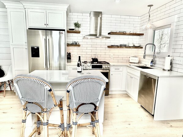 A modern kitchen with white cabinetry, a stainless steel refrigerator, a gas stove, and a dishwasher. There are two wicker chairs at a white table.
