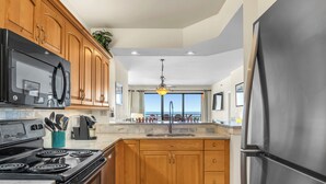Une cuisine moderne avec des meubles en bois, un réfrigérateur en acier inoxydable, une cuisinière noire et une vue sur l’océan grâce à de grandes fenêtres.