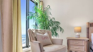 Une pièce avec une grande fenêtre, un fauteuil moelleux agrémenté d’un coussin en forme de cœur, une petite table de chevet avec une lampe et une plante en pot.