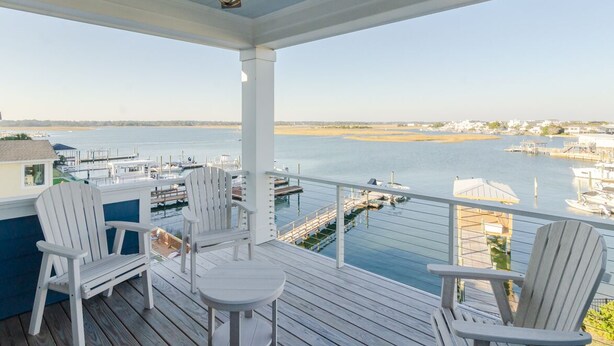 A covered deck with white Adirondack chairs, a ceiling fan, and a view of a marina with boats and a distant shoreline. - Sound front home w boat slip! New for 2026 pool! (Wrightsville Beach)
