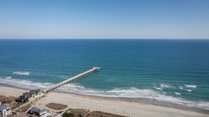 A coastal view with a long pier extending into the sea, a sandy beach, and buildings near the shore. - Sound front home w boat slip! New for 2026 pool! (Wrightsville Beach)