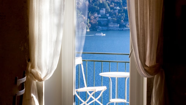 Un balcón con sillas y una mesa blancos, con vista a una masa de agua donde se ve un barco y edificios al fondo.