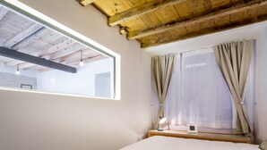 Un dormitorio con techo de madera, un ventanal con cortinas, una mesita de noche con lámpara y una cama.