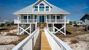 Une maison située en bord de mer, dotée d’une terrasse en bois qui mène à un porche abrité, et entourée de dunes de sable et d’autres maisons.