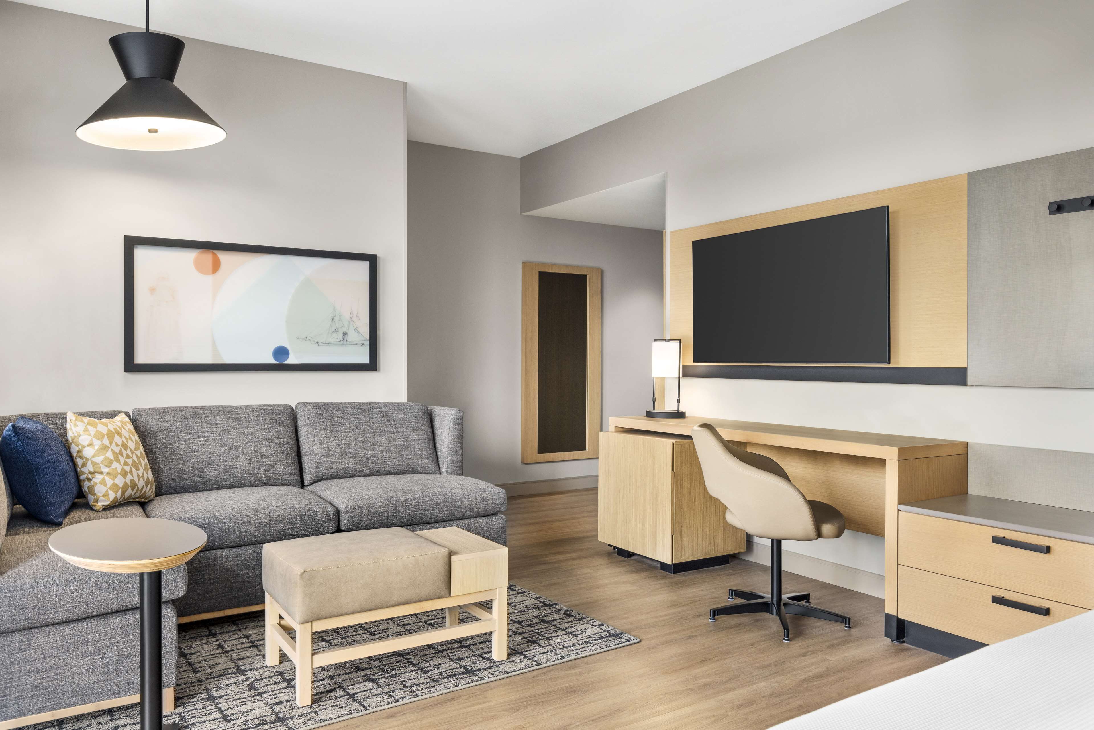 A modern hotel room with a grey sofa, a small round table, a wooden desk with a chair, a large flat-screen TV, and a framed abstract artwork on the wall.