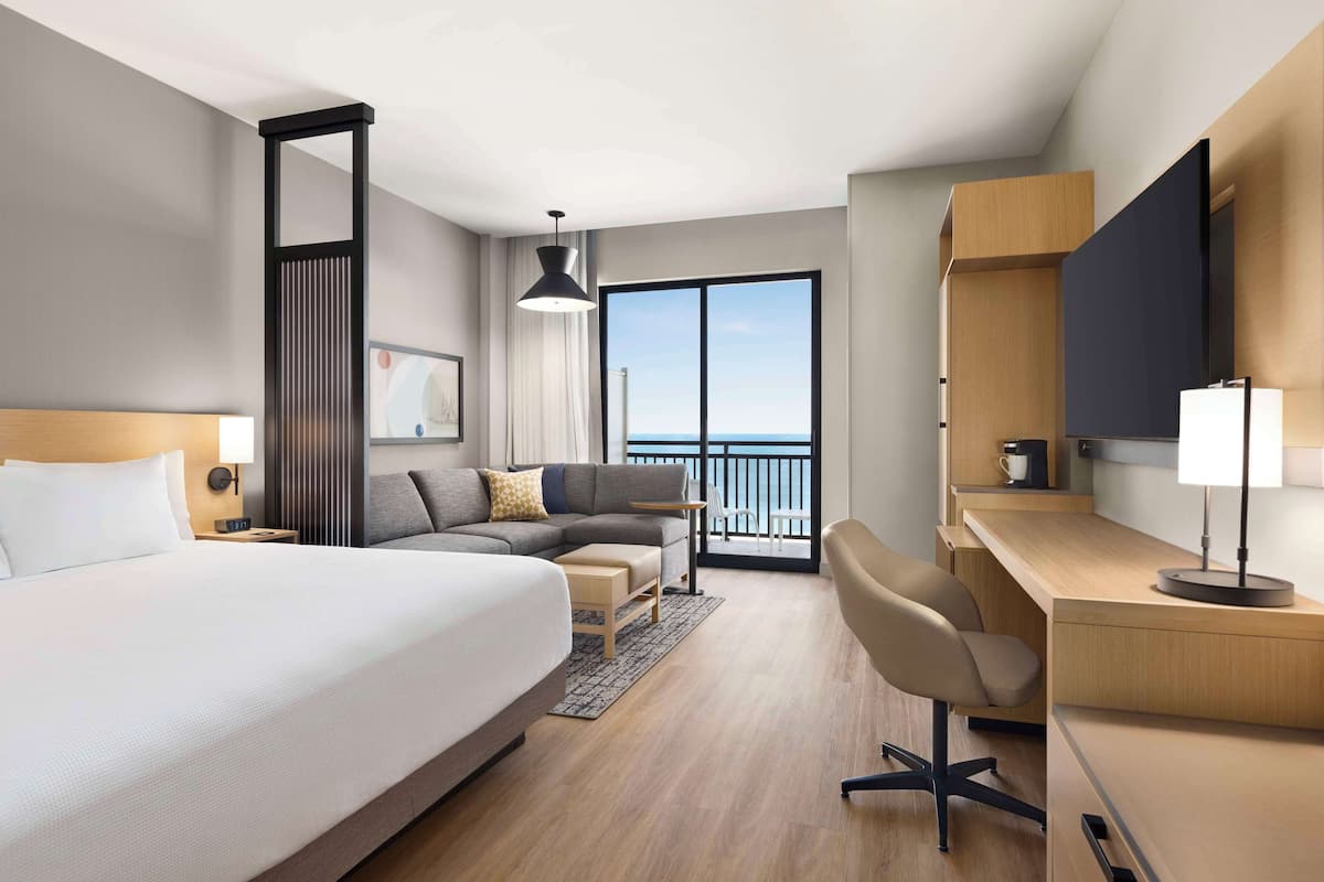 A modern hotel room with a large bed, a desk, a chair, a sofa, a TV, and a balcony with a view.