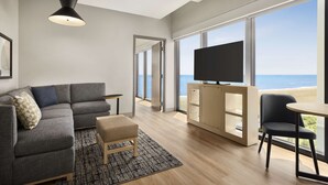 A modern living room with a gray sectional sofa, a small ottoman, a black chair, a wooden coffee table, a TV on a wooden cabinet, and large windows offering a view of the beach. - Hyatt Place Virginia Beach / Oceanfront (Virginia Beach)