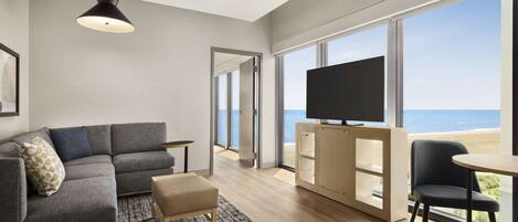 A modern living room with a gray sectional sofa, a small ottoman, a black chair, a wooden coffee table, a TV on a wooden cabinet, and large windows offering a view of the beach.