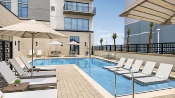 A rooftop pool area with lounge chairs and umbrellas, surrounded by a modern building with balconies and large windows.