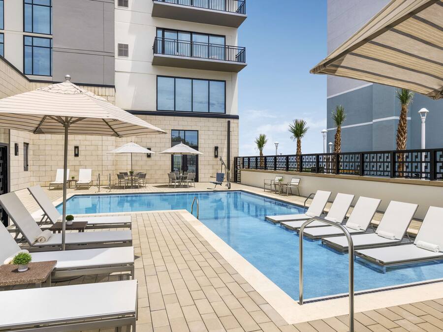 A rooftop pool area with lounge chairs and umbrellas, surrounded by a modern building with balconies and large windows.