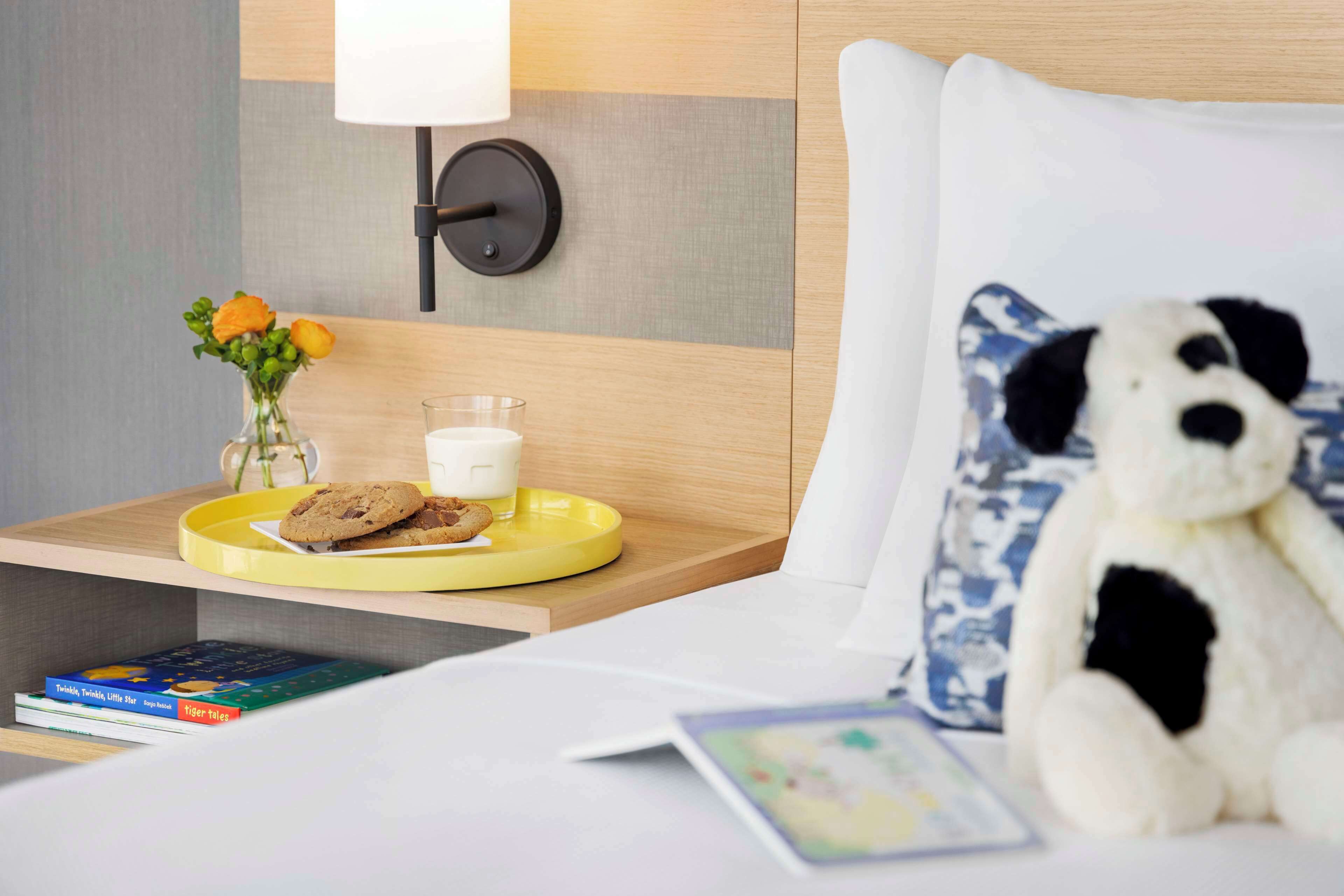 Une chambre d’hôtel bien rangée, avec un lit, une table de chevet sur laquelle se trouve une lampe, un ours en peluche, un vase rempli de fleurs et un plateau avec des biscuits et un verre de lait.