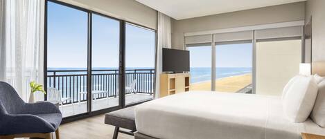 Une chambre d’hôtel avec un grand lit, une télévision, un balcon donnant sur la plage et un coin salon avec un fauteuil et une plante.