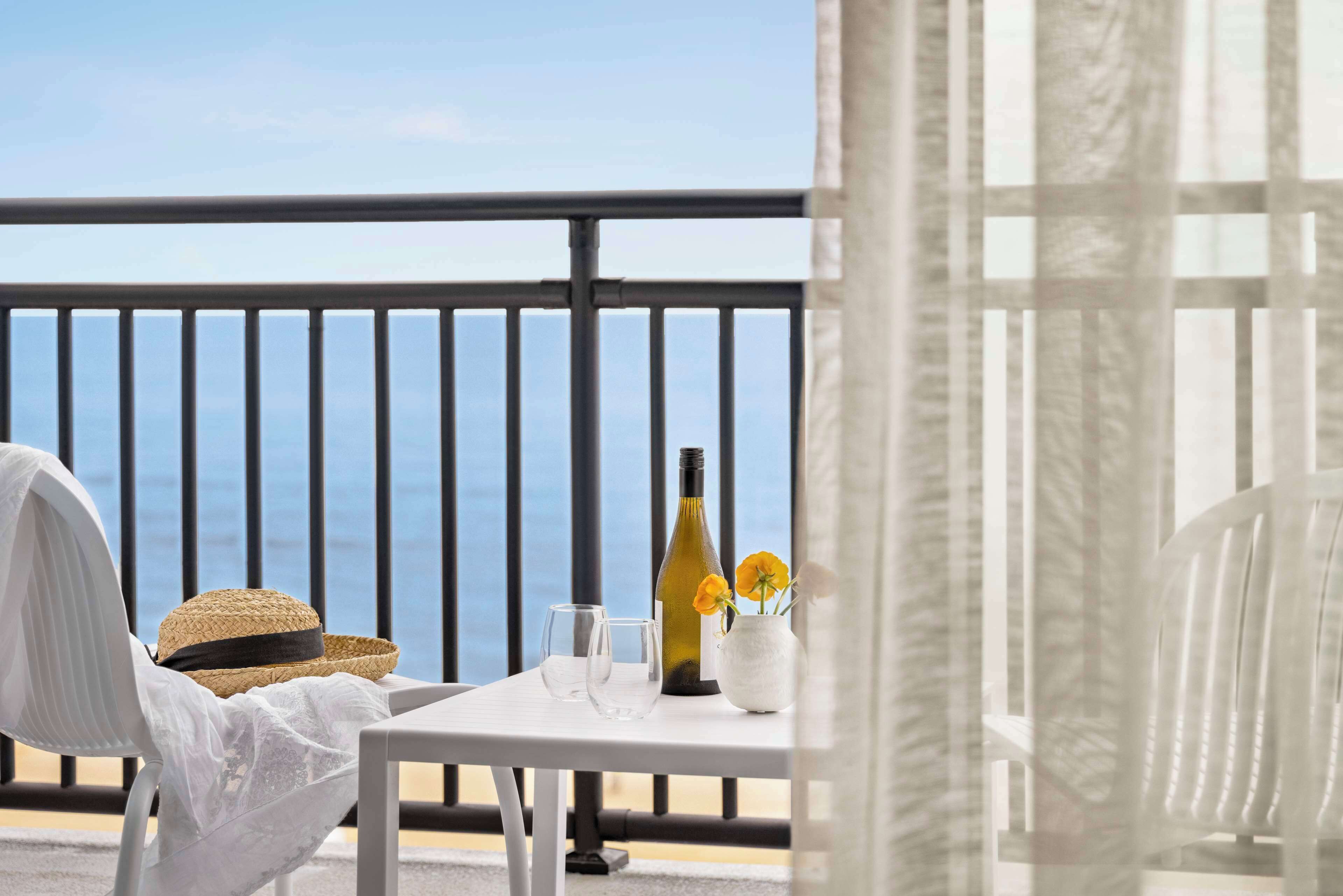 A balcony with a white chair, a table with a bottle and glasses, and a straw hat.