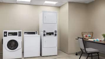 Un cuarto de lavandería con tres lavadoras y secadoras apiladas, una silla de oficina gris y una cesta tejida sobre una encimera.