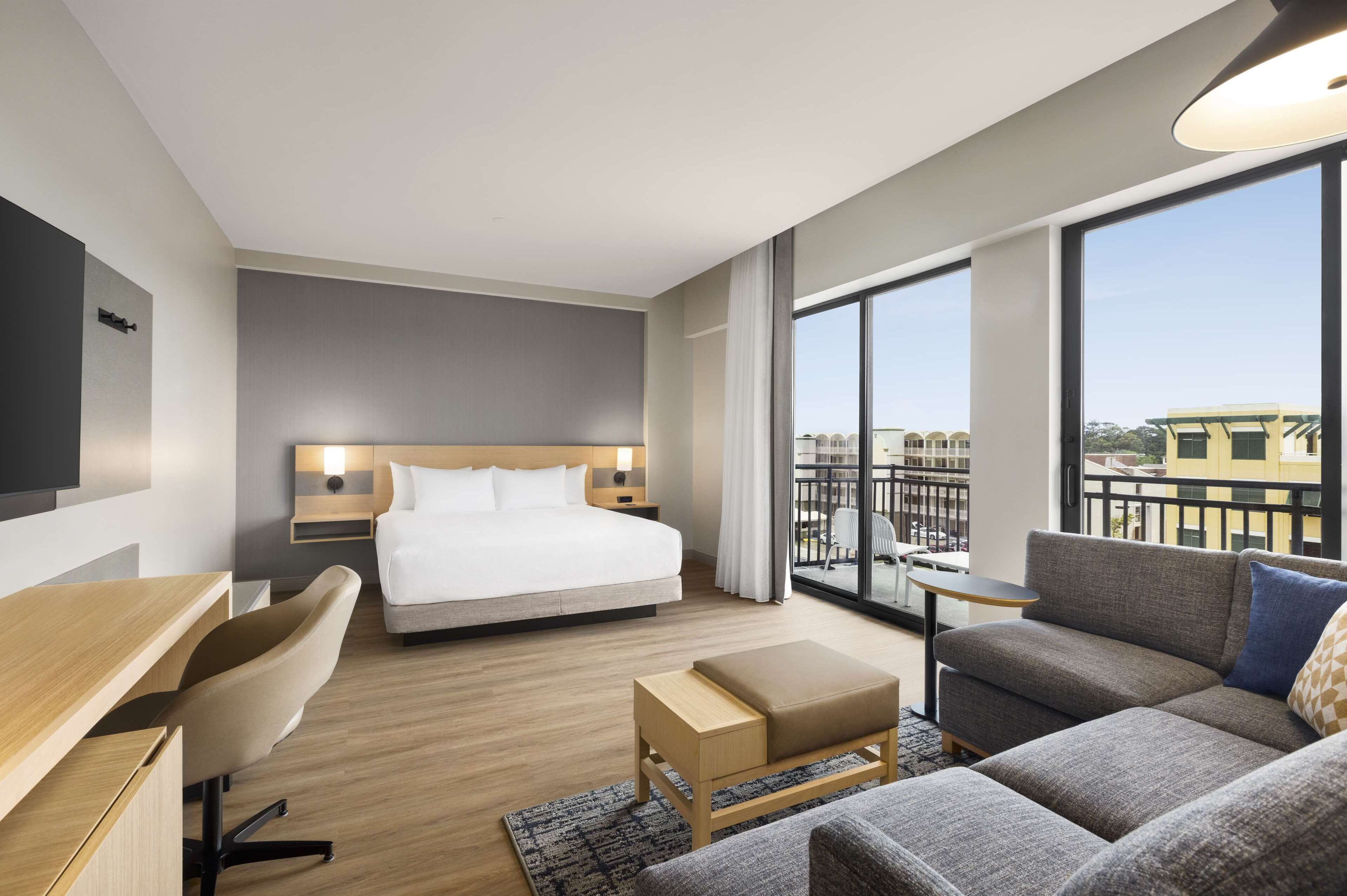 Habitación de hotel moderna con una cama grande, un sofá, un escritorio y un balcón con vistas a edificios.