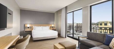 A modern hotel room with a large bed, a sofa, a desk, and a balcony with a view of buildings.
