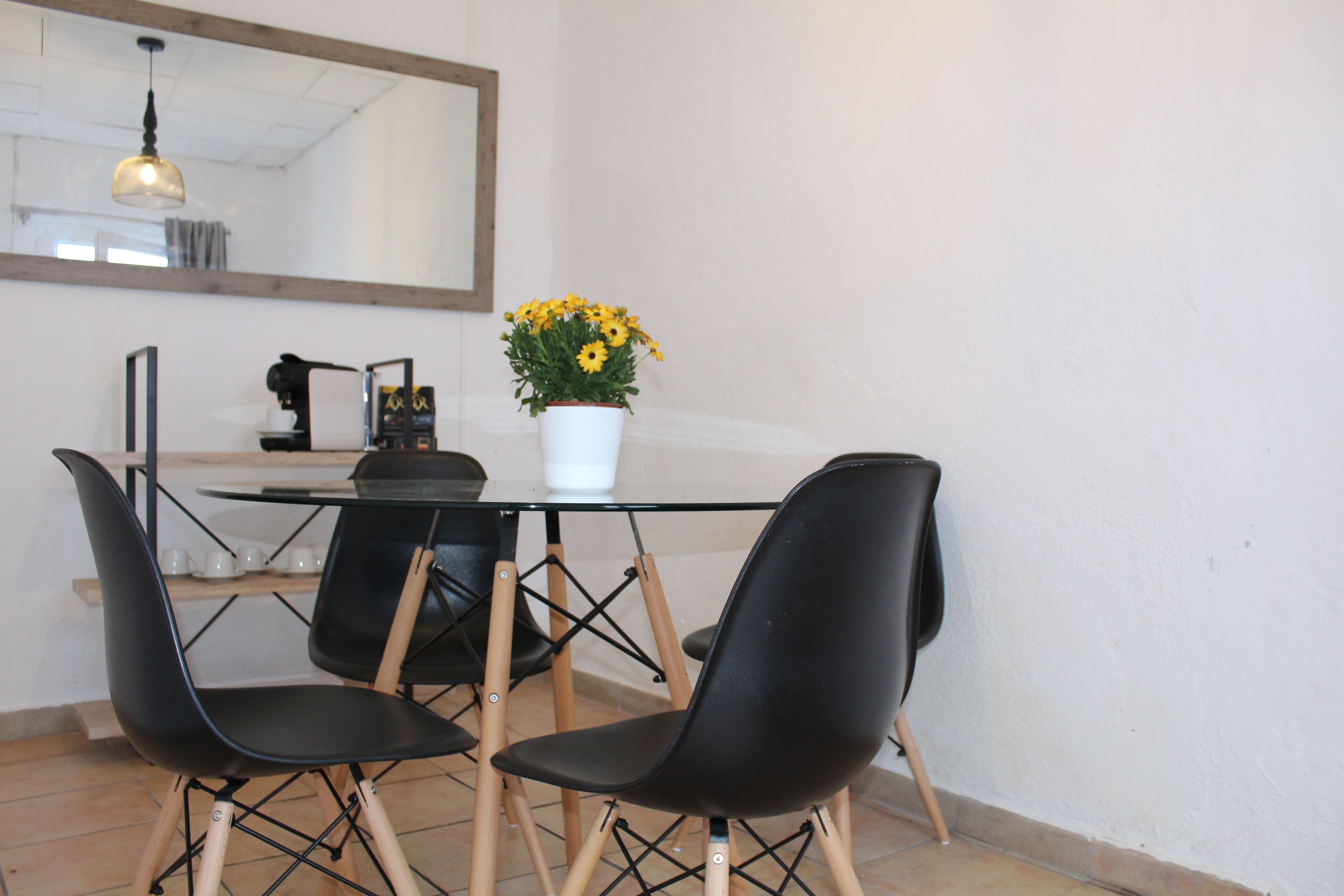 Un comedor con mesa de vidrio, sillas negras, un jarrón blanco con flores amarillas y una cafetera.