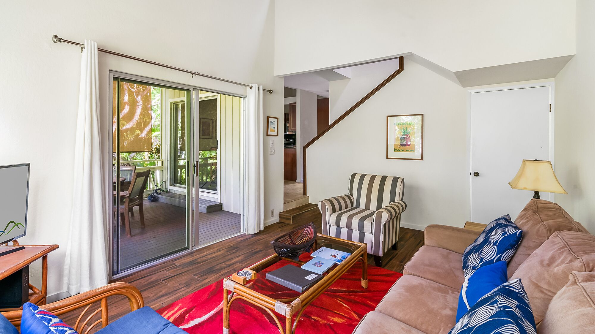 Una sala de estar con un sofá, un sillón a rayas, una mesa de centro con libros, un televisor y un ventanal corredero que da a un área exterior.