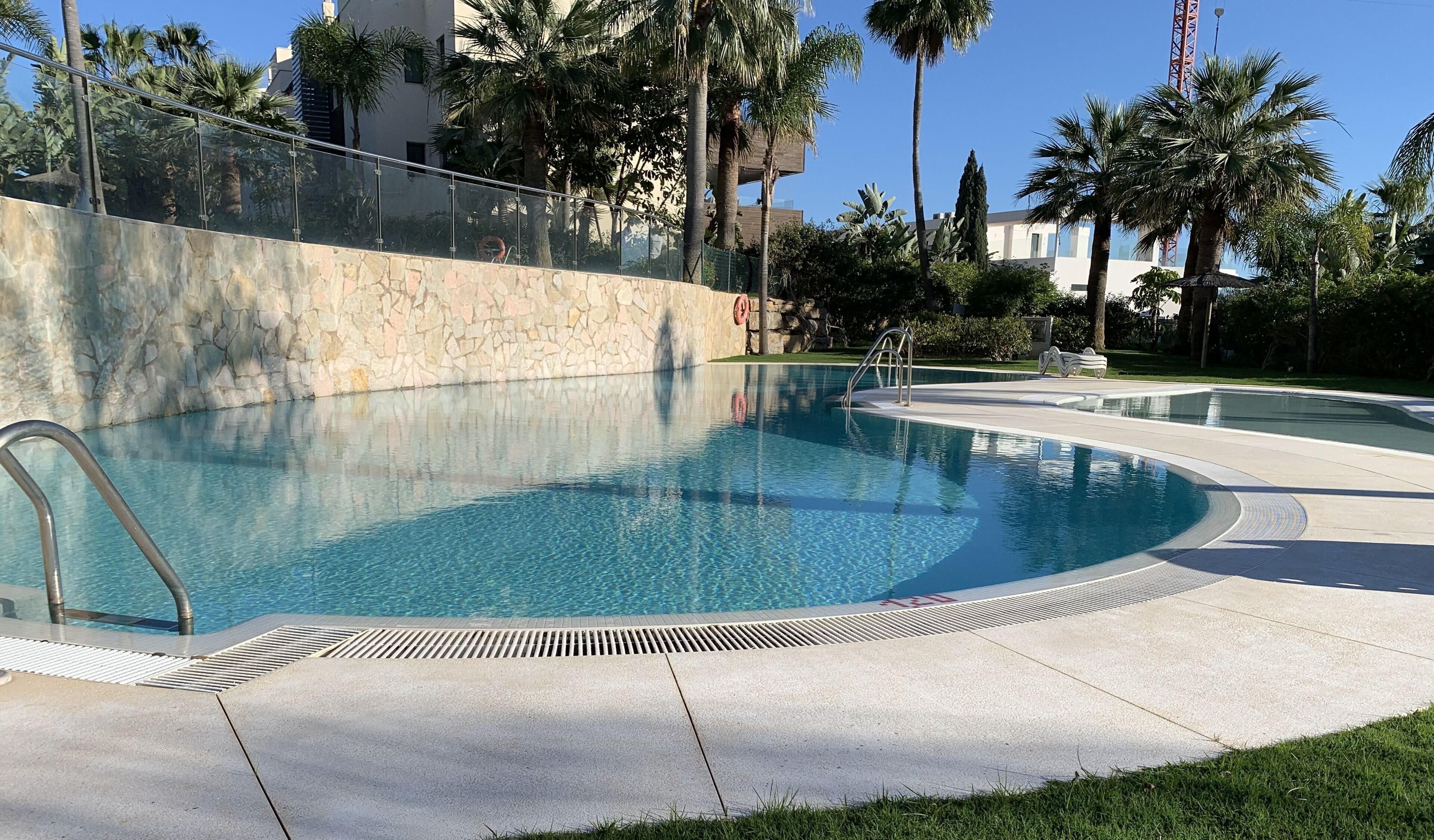 Une piscine bordée d’un mur en pierre, avec une rambarde en métal et des palmiers en arrière-plan.