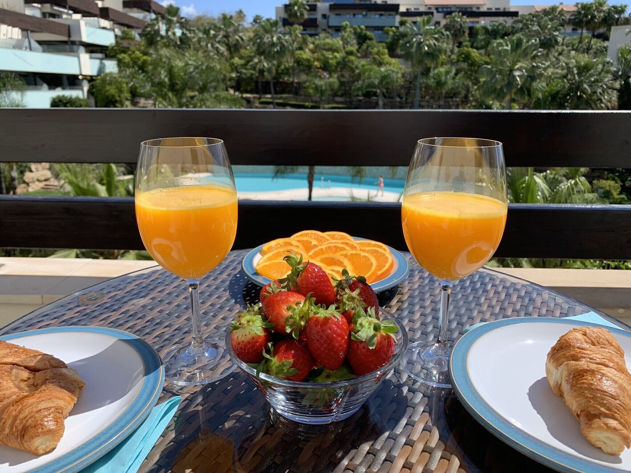 A table with two glasses of orange juice, a bowl of strawberries, and a croissant on a balcony overlooking a pool and palm trees.