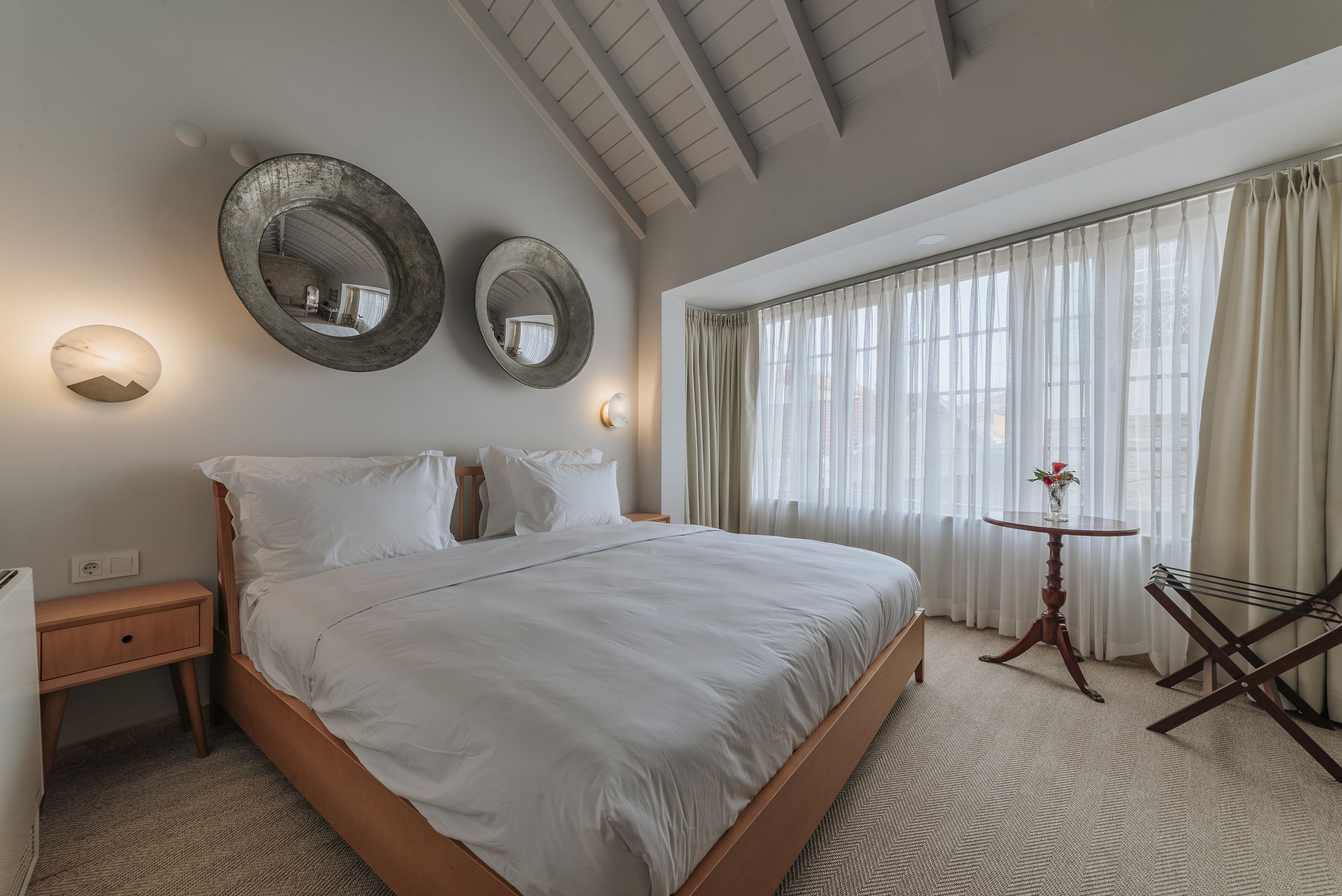 A hotel room with a large bed, two wall-mounted mirrors, a bedside table, a small round table with a vase, and a window with sheer curtains.
