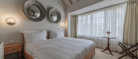 A hotel room with a large bed, two wall-mounted mirrors, a bedside table, a small round table with a vase, and a window with sheer curtains.