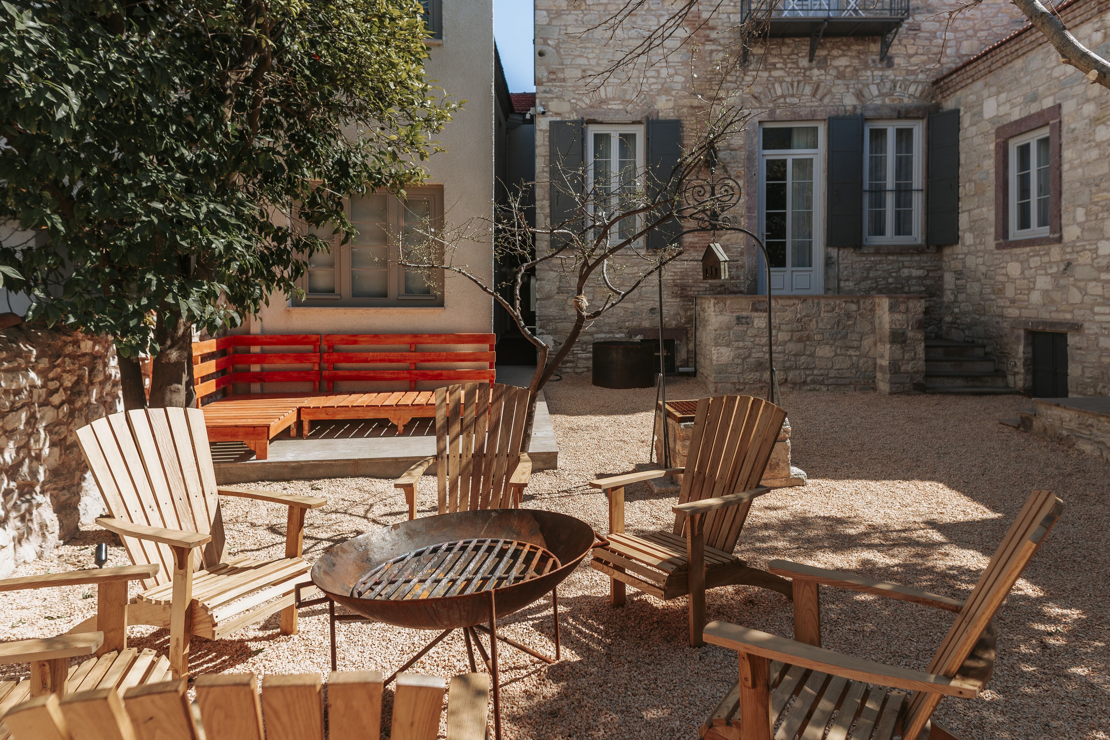 An outdoor seating area with wooden chairs and a metal fire pit, surrounded by stone buildings and a tree.