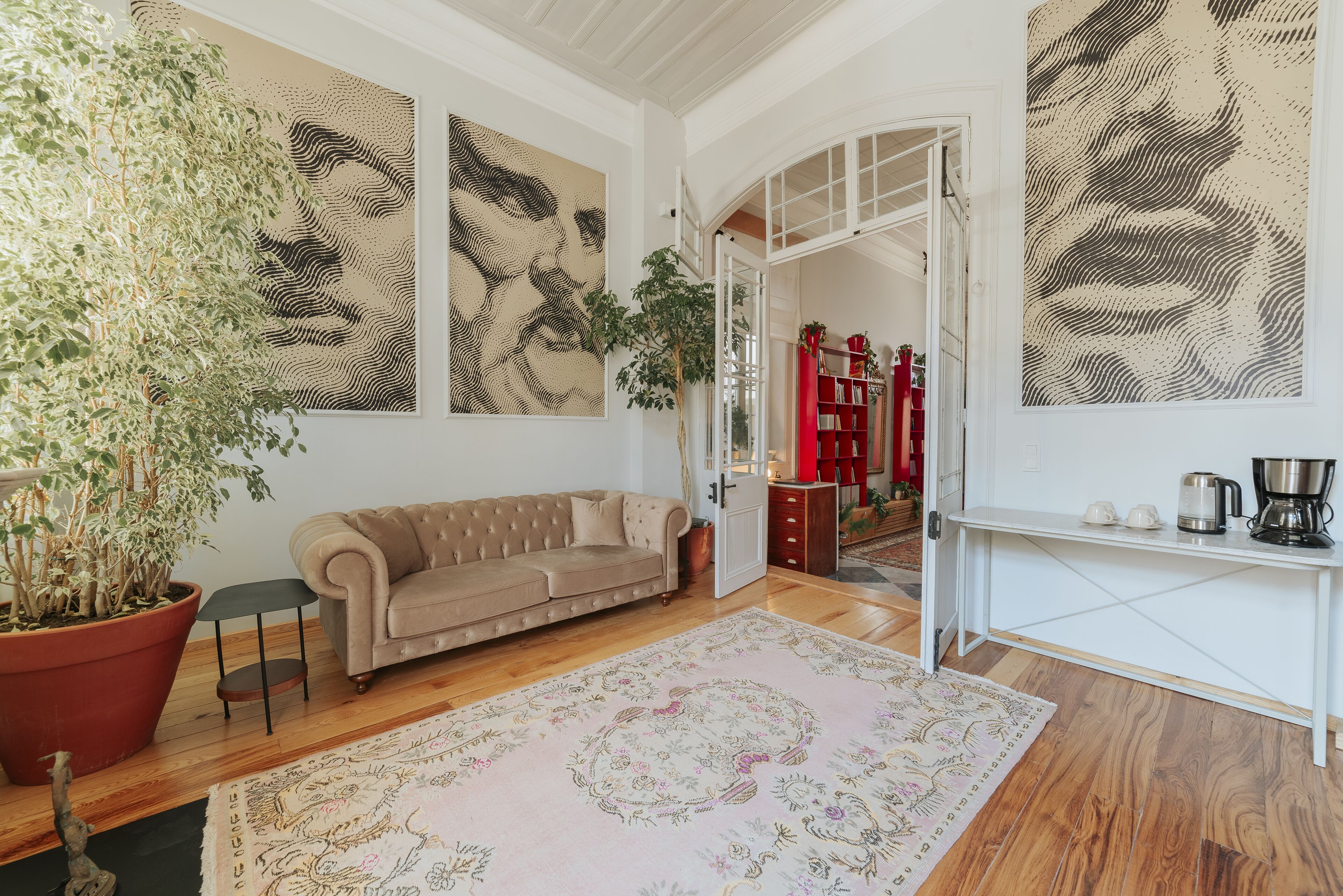 A living room with a beige sofa, a large potted plant, and a patterned rug. There are abstract wall art pieces and a white console table with a kettle.
