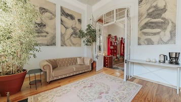 Un salon avec un canapé beige, une grande plante en pot et un tapis à motifs. On y trouve des œuvres d’art murales abstraites et une console blanche sur laquelle est posée une bouilloire.