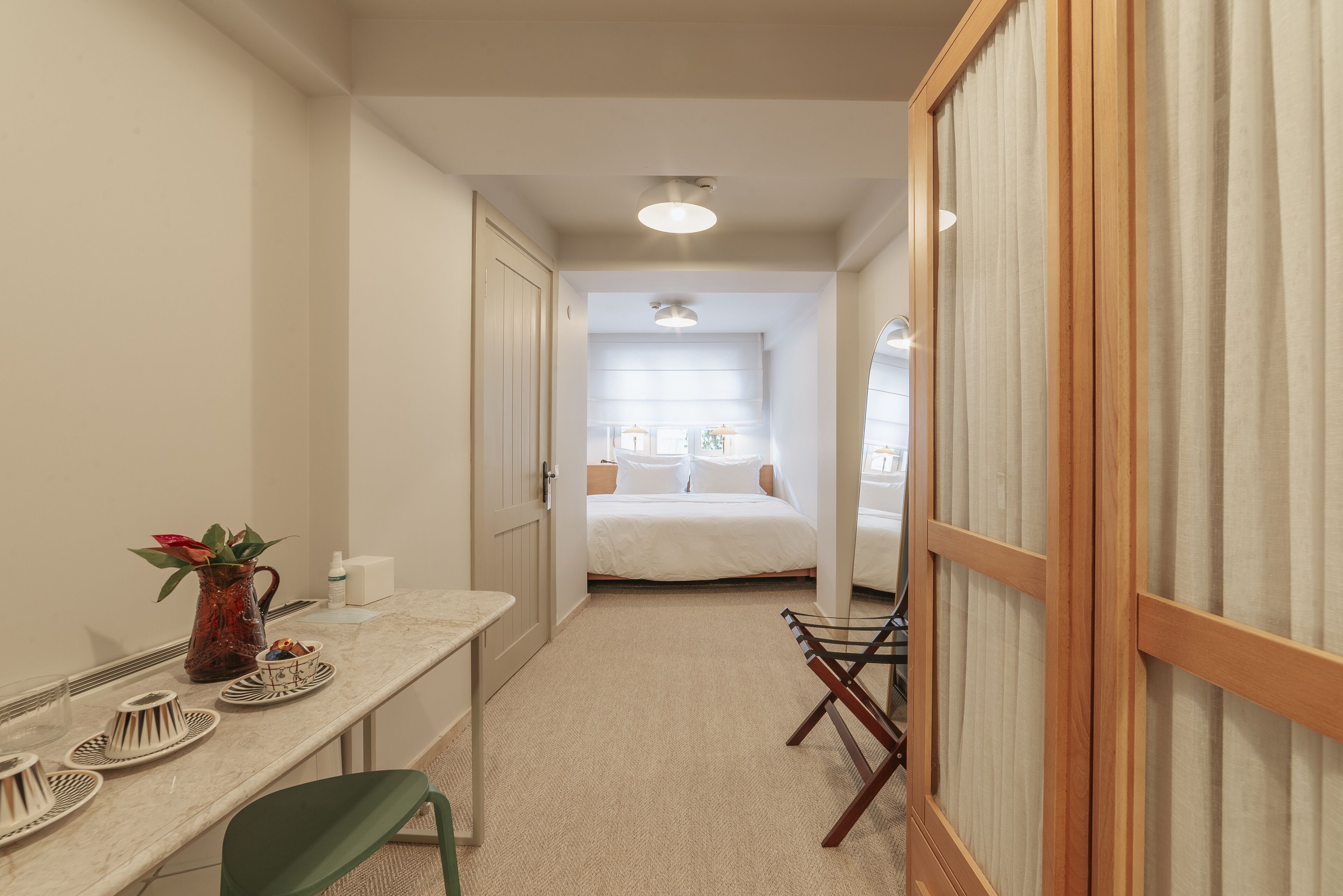 Un couloir étroit mène à une chambre à coucher qui contient un lit, un bureau avec une chaise et une petite table sur laquelle se trouvent un vase et des assiettes.