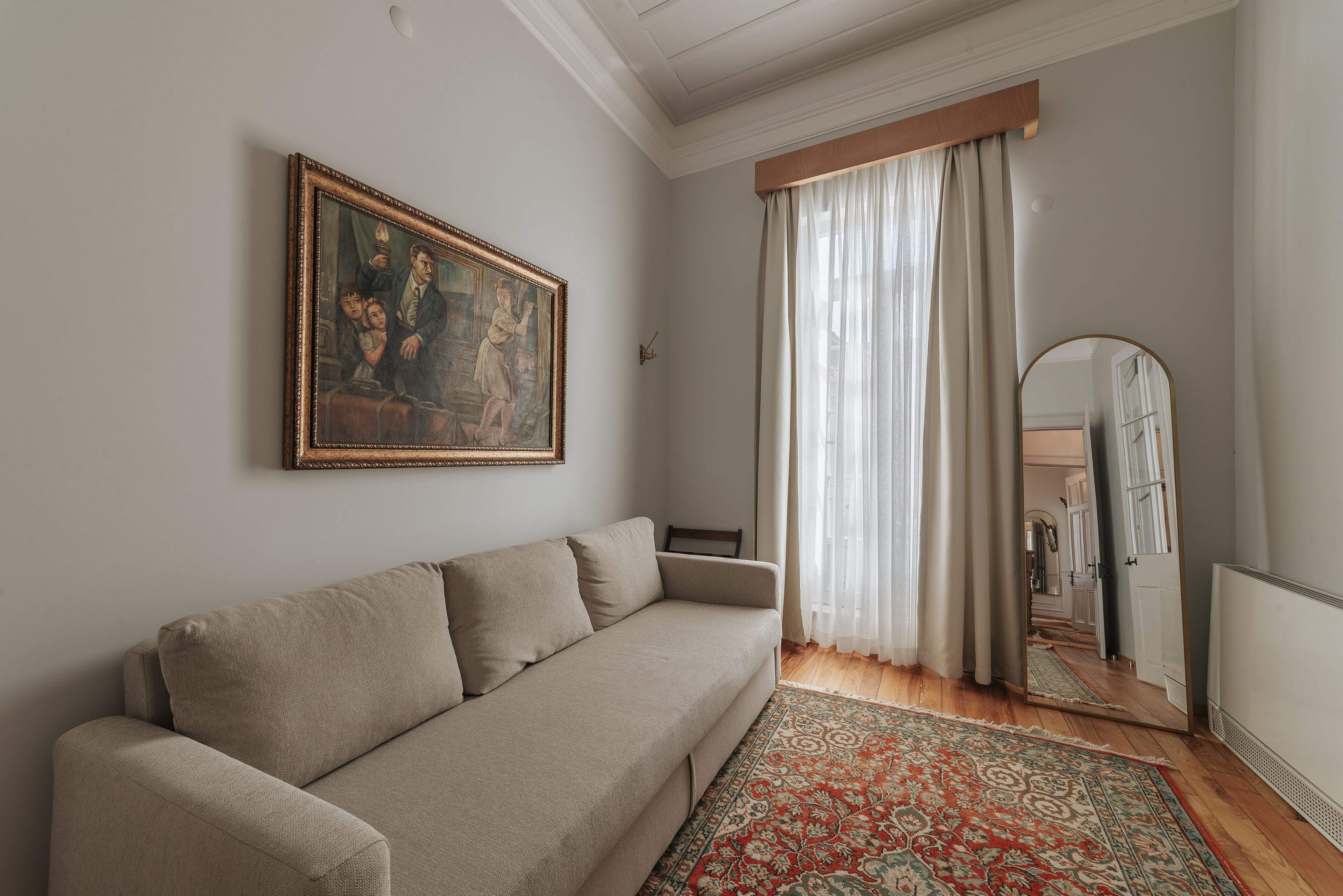 Una sala de estar con un sofá beige, un cuadro en la pared, un ventanal con cortinas traslúcidas y un espejo que refleja la habitación.