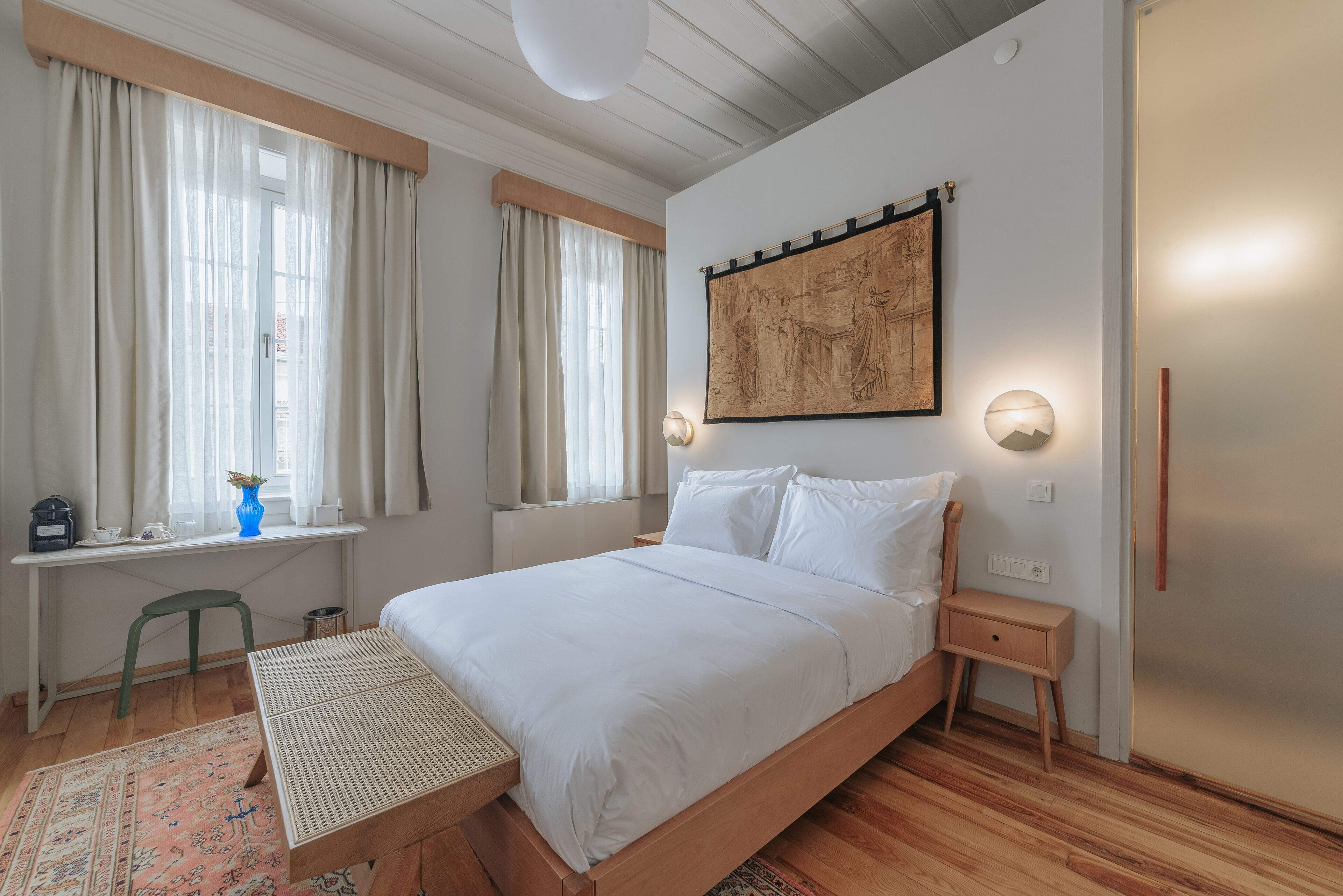 Un dormitorio con cama, mesitas de noche, un escritorio con silla, una ventana con cortinas y una obra de arte colgada en la pared.