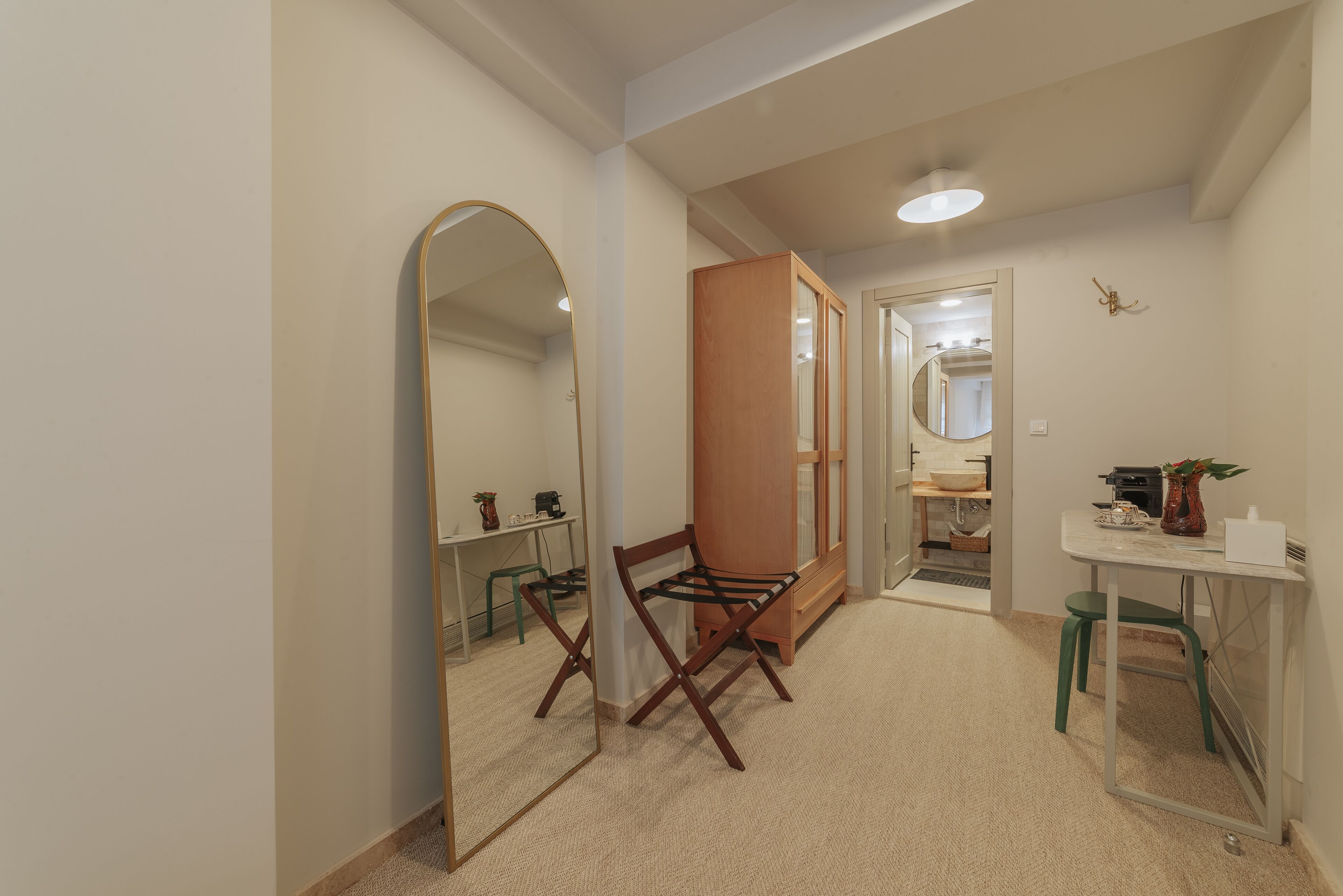 A room with a large mirror, a wooden wardrobe, a desk with a chair, and a small table with a vase.