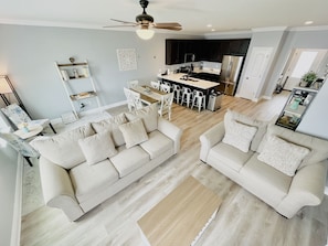 Un salon moderne avec deux canapés beige, un coin repas avec des chaises blanches et une cuisine dotée de meubles sombres et d’appareils électroménagers en acier inoxydable.
