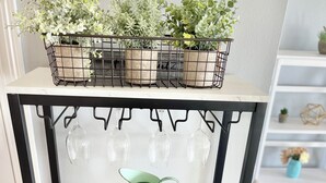 Une cuisine dotée d’un plan de travail en marbre, d’un présentoir en métal noir sur lequel sont disposées des plantes en pot, de verres à vin, d’un arrosoir vert, d’un pot de miel et d’un plateau en bois contenant des cure-dents.
