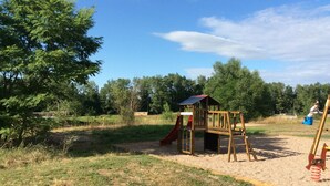 Un parque infantil con una estructura de madera que incluye un tobogán y columpios, sobre una zona de arena con césped y árboles al fondo.
