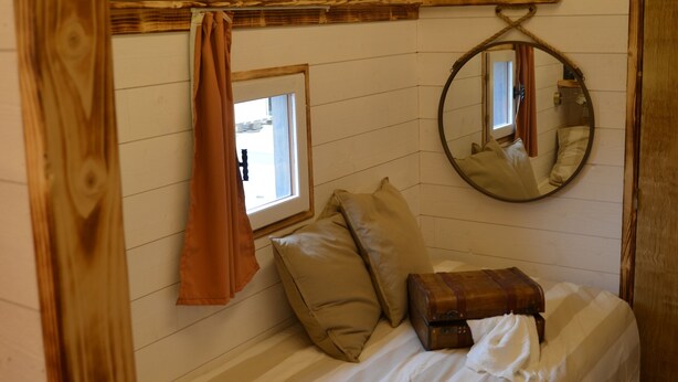 Un dormitorio rústico con techo de madera, una cama individual con ropa de cama a rayas, una ventana pequeña y un espejo redondo en la pared.