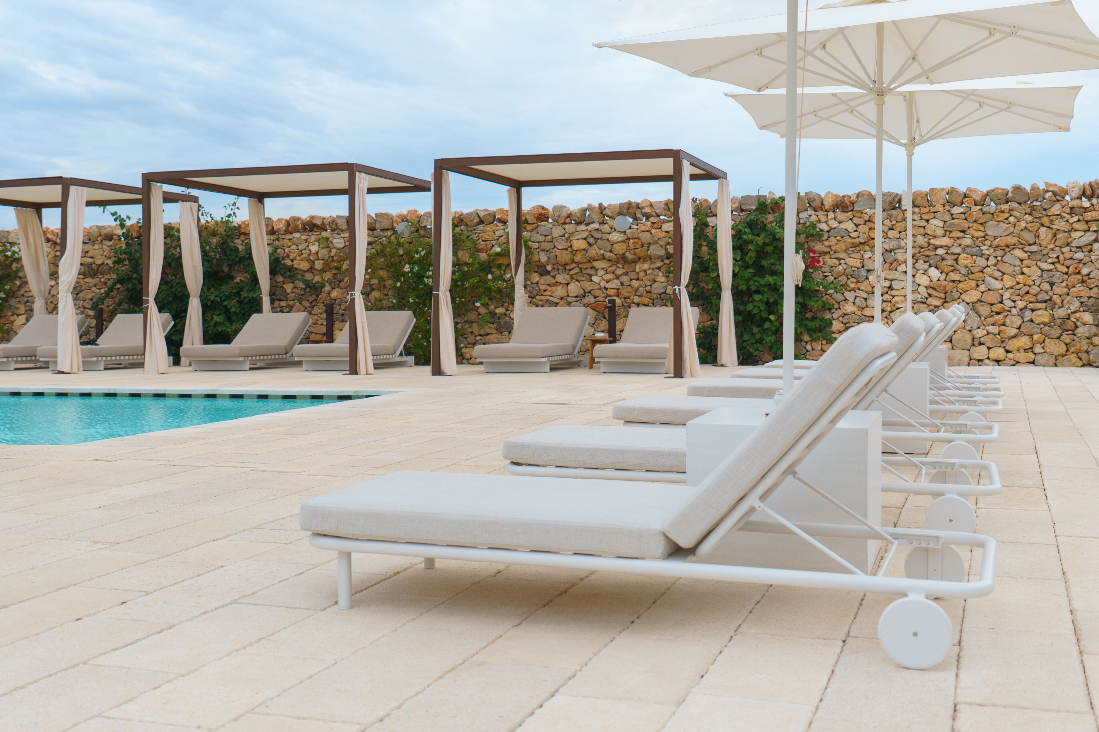 A poolside area with lounge chairs and umbrellas, a stone wall, and a clear sky.