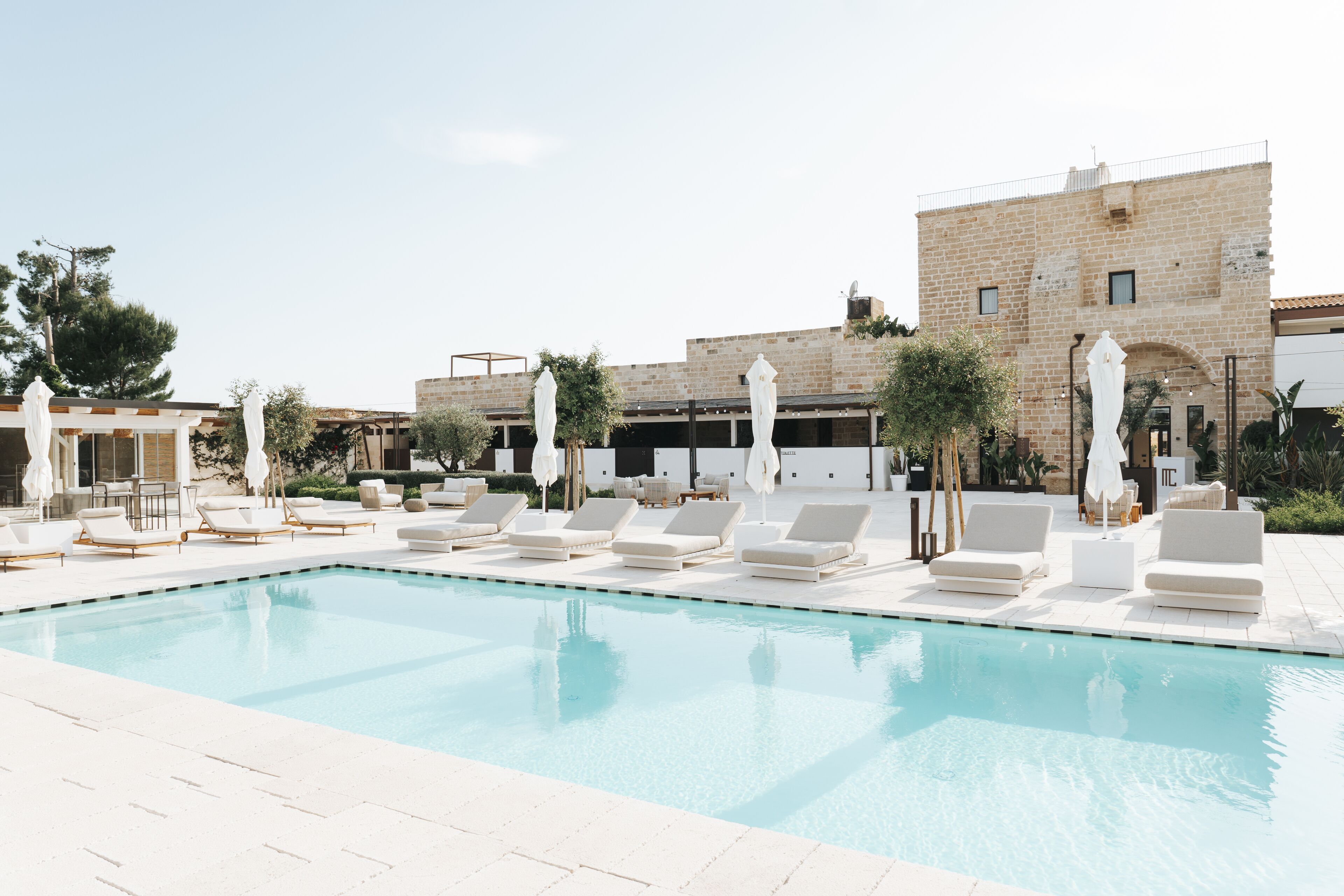 Zona de piscina con sillones reclinables y sombrillas, un edificio de piedra al fondo y un cielo despejado.