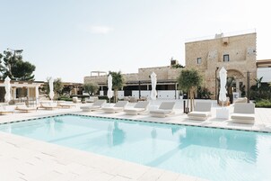 Zona de piscina con sillones reclinables y sombrillas, un edificio de piedra al fondo y un cielo despejado.