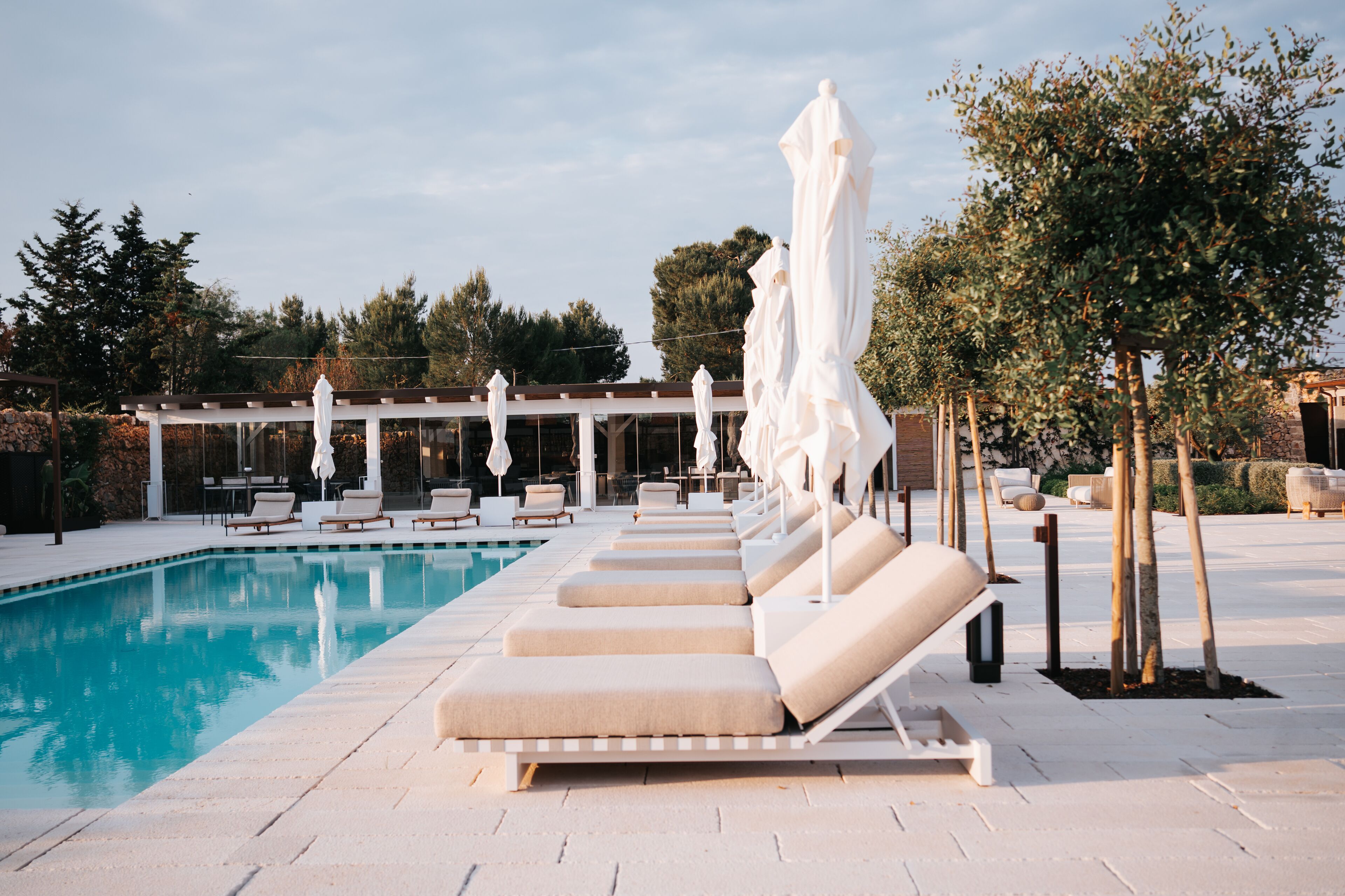 A pool area with lounge chairs and umbrellas, surrounded by trees and a building in the background.