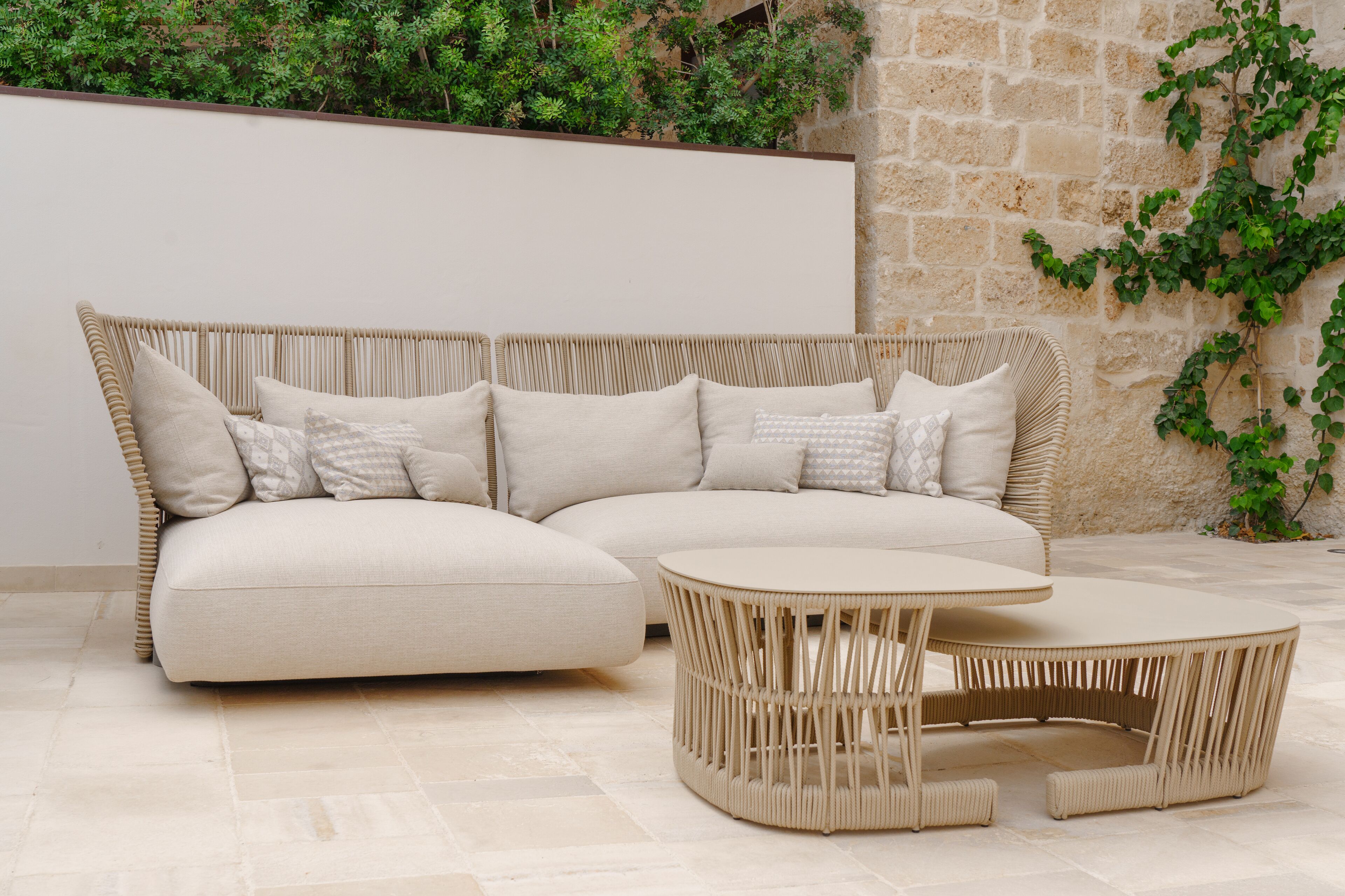 Un sofá seccional color beige con varios cojines, una mesa de centro redonda trenzada y una pared de piedra con plantas.