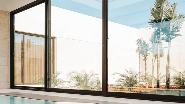 A modern indoor swimming pool with large glass doors leading to a view of palm trees and desert landscape. - Masseria Corsano (Nardò)