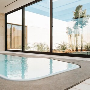A modern indoor swimming pool with large glass doors leading to a view of palm trees and desert landscape. - Masseria Corsano (Nardò)