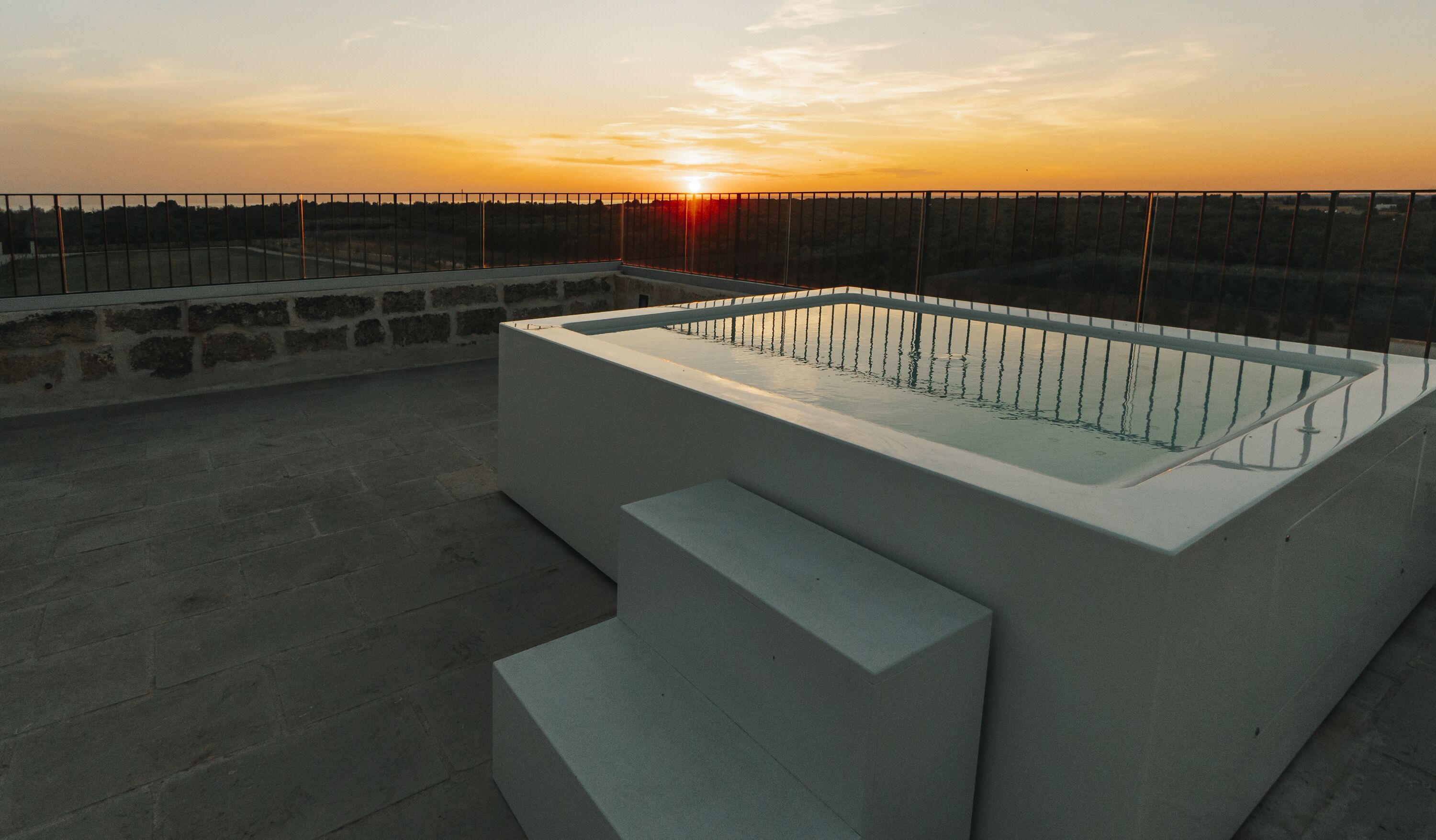Une piscine extérieure moderne, dotée d’une paroi en verre et de marches, qui se détache sur le décor d’un coucher de soleil.