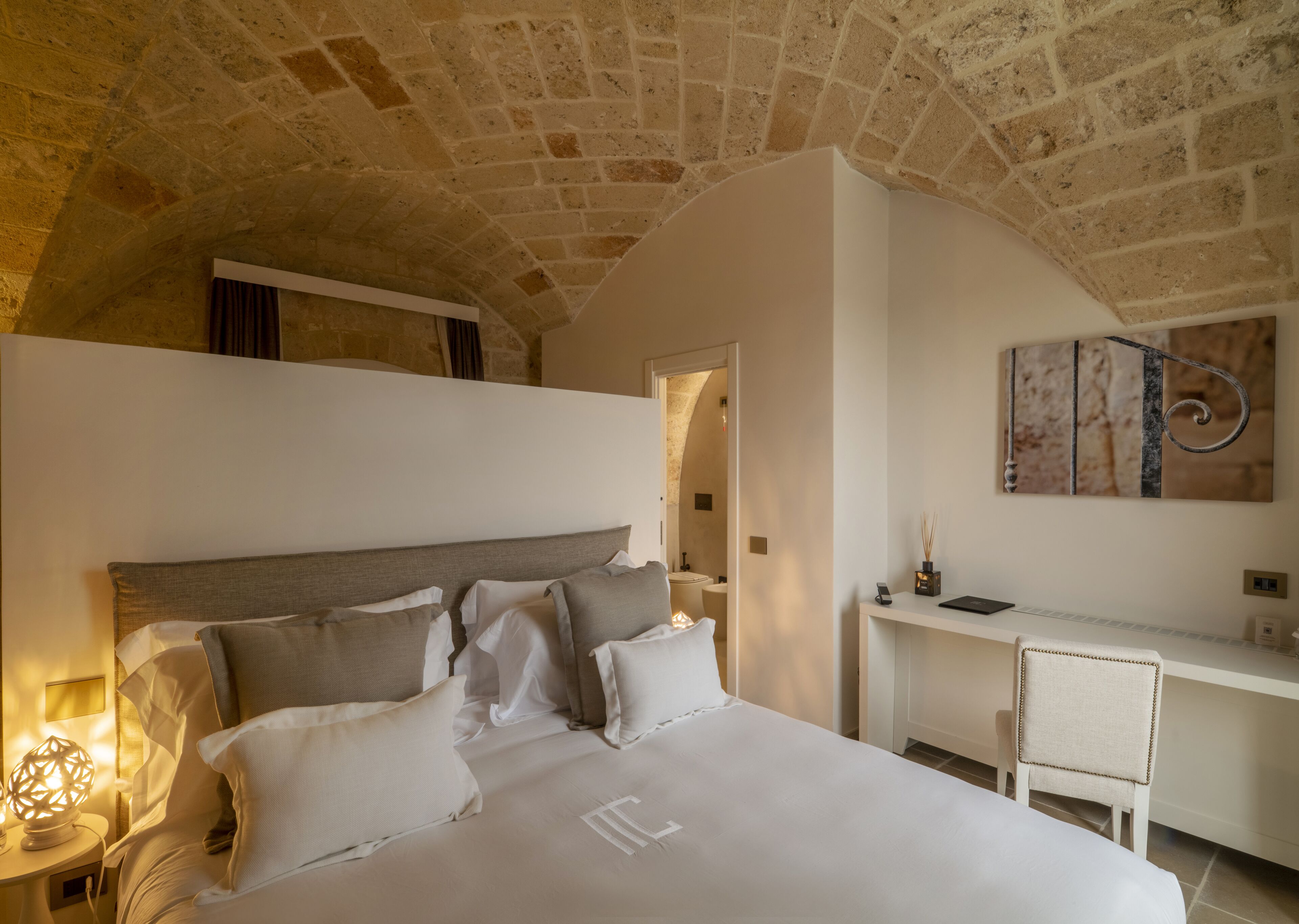 Un dormitorio con techo de piedra, una cama con ropa de cama blanca, un escritorio y un cuadro en la pared.